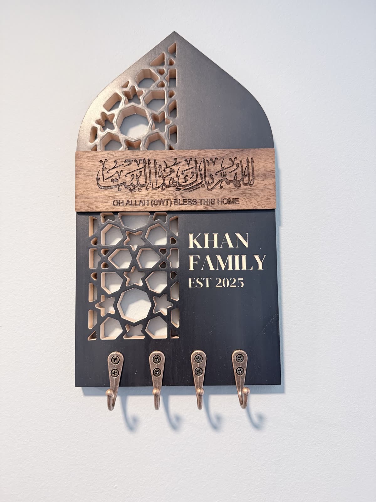 An arched key holder on a white wall with a walnut calligraphy band, lattice cut-outs, a family name and four hooks
