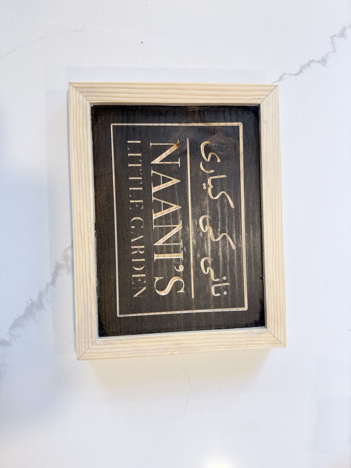 A personalized name sign with Arabic and English lettering on a dark painted backer in a pale wood frame